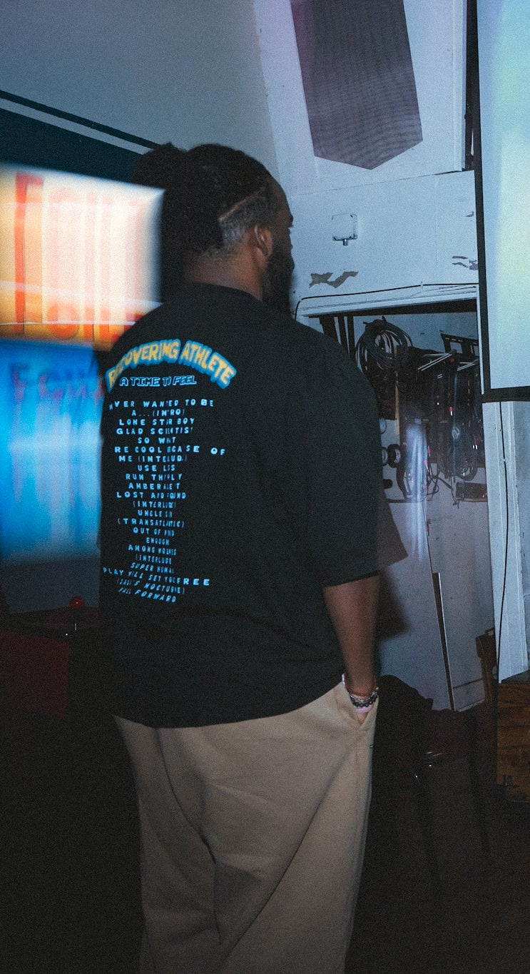 Ryan The Son wearing the Recovering Athlete tee at a live show, back graphic visible against a projection screen