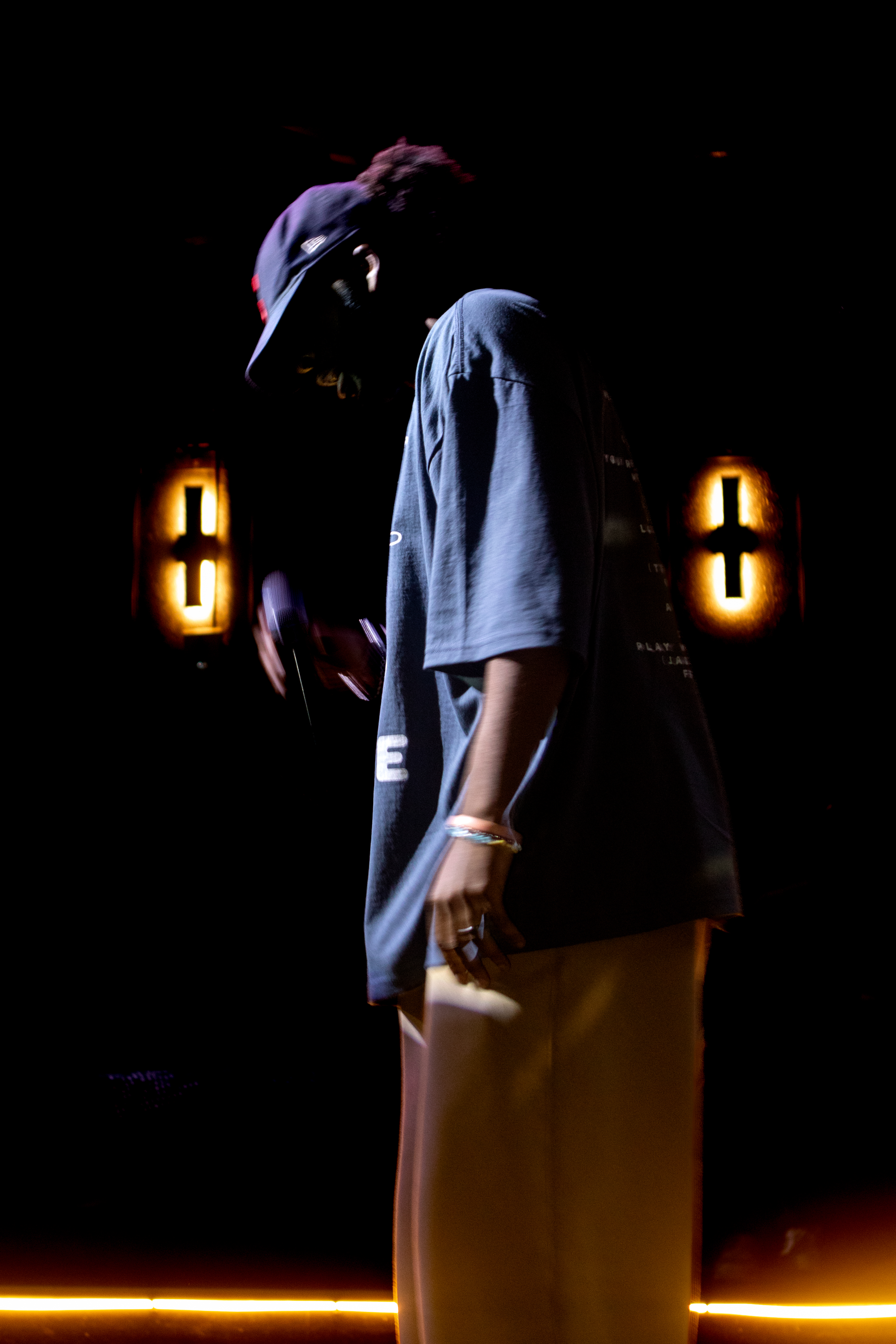 Ryan The Son in profile on stage, PWSYF tee silhouetted by warm stage lights